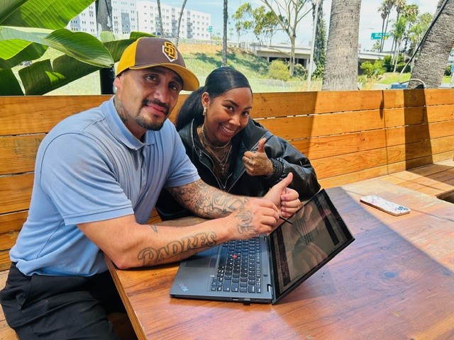 Two people sitting at a table with a laptop, one is giving a thumbs up